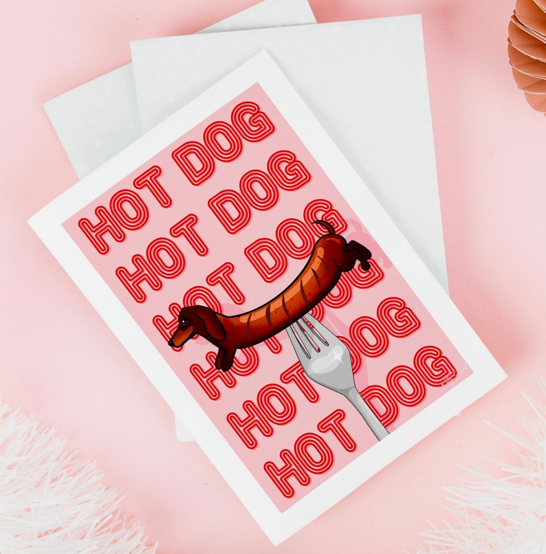 Printable pink & red dog art print. Soft pink background with red text in the style of neon lights reading HOT DOG. in the centre of the art print is a dachshund in the shape of a sausage on the end of a silver fork in a playful style on pink table