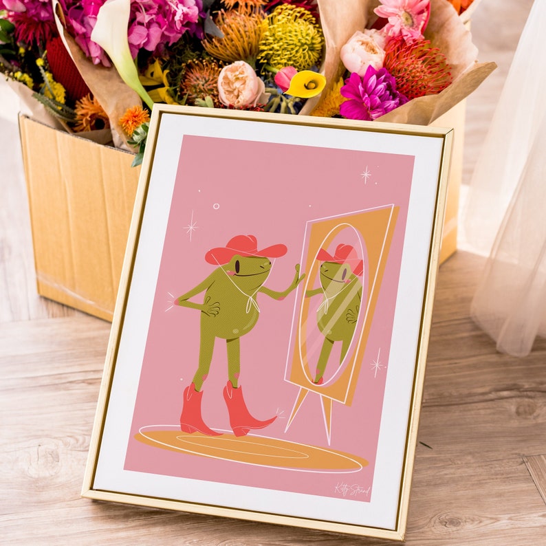 pink and green cowboy frog art print of a cartoon style frog wearing a pink cowgirl hat and pink cowboy boots looking into a vintage yellow mirror. Print features a soft pink background and sits in a gold frame resting against a box of flowers