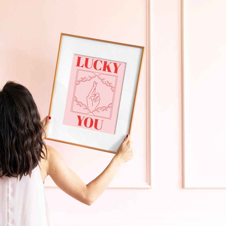 women in a pink living room holding a golden frame. Art in frame is a lucky you fingers crossed art print in pink and red with line art and retro lettering