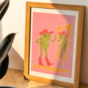 Art print of a frog wearing western boots and a cow girl hat looking in a mirror. Print is in a wooden frame next to a house plant. Both are bathed in sunlight