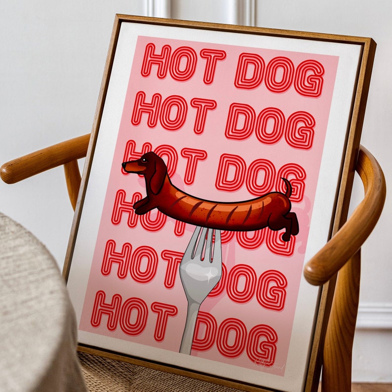 Printable pink & red dog art print. Soft pink background with red text in the style of neon lights reading HOT DOG. in the centre of the art print is a dachshund in the shape of a sausage on the end of a silver fork. Print rests in mid century chair