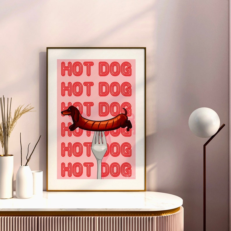Printable pink & red dog art print. Soft pink background with red text in the style of neon lights reading HOT DOG. in the centre of the art print is a dachshund in the shape of a sausage on the end of a silver fork in a playful style in wood frame