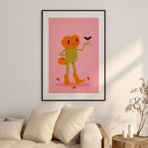 howdy halloween frog art print in pink, green and orange hangs on wall. UPS style cartoon illustration of a frog with a pumpkin head wearing orange cowboy boots and carrying a pumpkin bag. next to him flies a little bat, leaves surround his feet