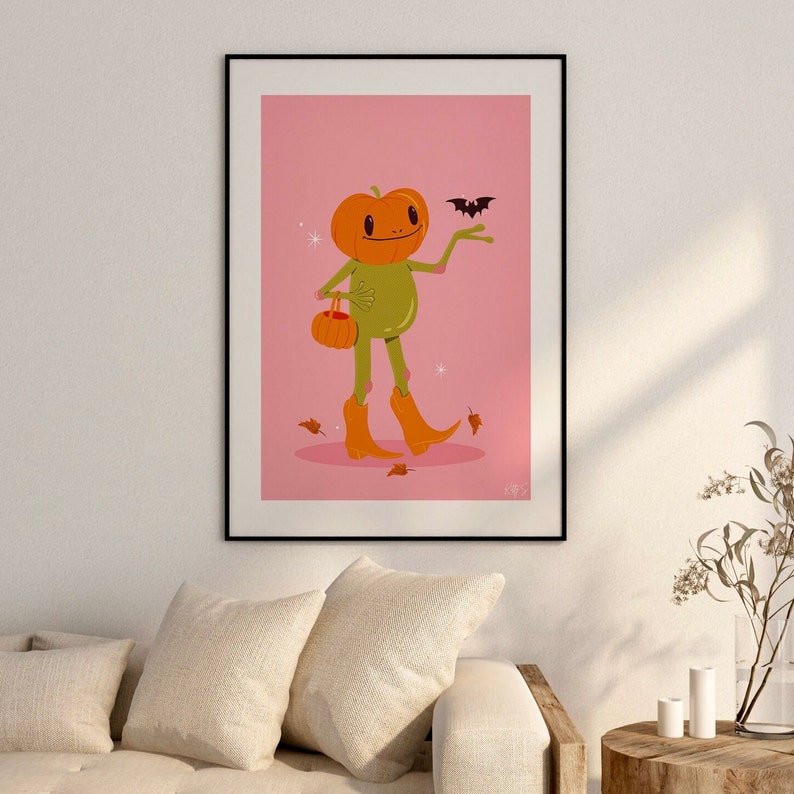 howdy halloween frog art print in pink, green and orange hangs on wall. UPS style cartoon illustration of a frog with a pumpkin head wearing orange cowboy boots and carrying a pumpkin bag. next to him flies a little bat, leaves surround his feet
