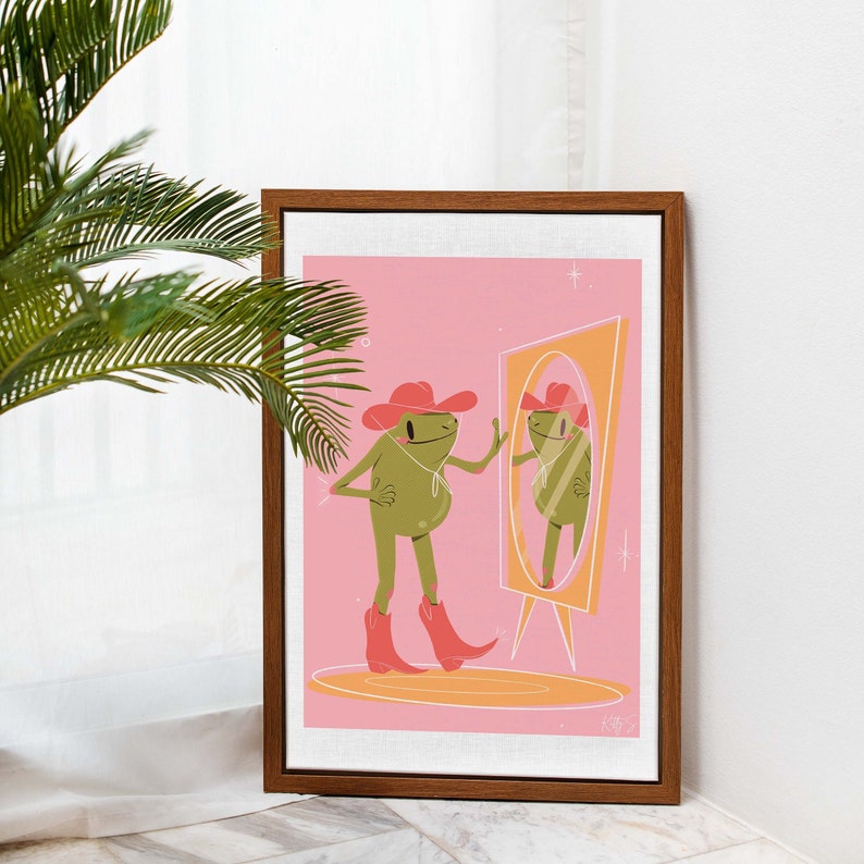 wooden frame leans against a white wall next to a tall green plant. Art print in the frame features a UPA style frog illustration. A Green smiling frog looks into a tall yellow mirror wearing pink cowgirl boots and a pink cowgirl hat