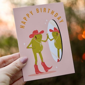 women holds cowboy frog happy birthday card. pink greetings card with a green frog in a cowboy hat and cowboy boots looking I the mirror and smiling. text on the card reads happy birthday in yellow lettering.