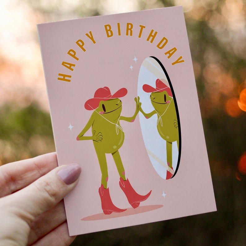 women holds cowboy frog happy birthday card. pink greetings card with a green frog in a cowboy hat and cowboy boots looking I the mirror and smiling. text on the card reads happy birthday in yellow lettering.