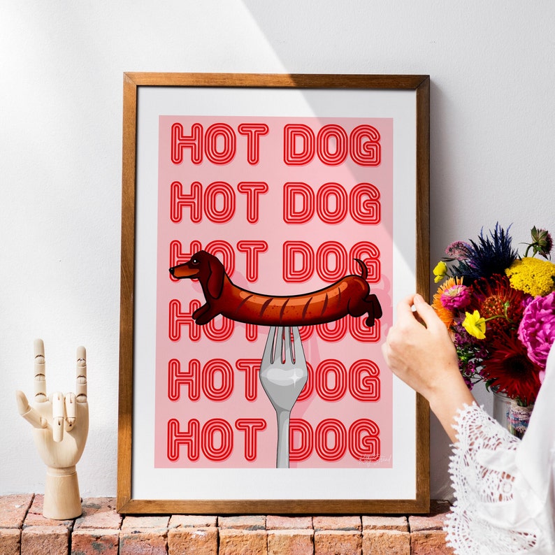 Printable pink & red dog art print. Soft pink background with red text in the style of neon lights reading HOT DOG. in the centre of the art print is a dachshund in the shape of a sausage on the end of a silver fork in a playful style in wood frame