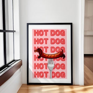 Printable pink & red dog art print. Soft pink background with red text in the style of neon lights reading HOT DOG. in the centre of the art print is a dachshund in the shape of a sausage on the end of a silver fork in a playful style in black frame