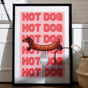 Printable pink & red dog art print. Soft pink background with red text in the style of neon lights reading HOT DOG. in the centre of the art print is a dachshund in the shape of a sausage on the end of a silver fork in a playful style in black frame