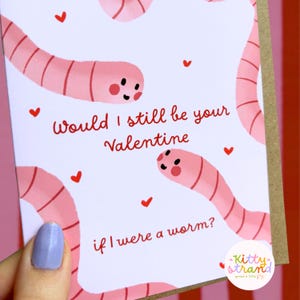 worm Valentine's Day card. pink Valentine's day card with an illustration of two worms surrounded by love hearts and text reading Would I still be your valentine if I were a worm? in red typography. Valentine's Day card for boyfriend, husband or partner held by a woman in front of a cosy background
