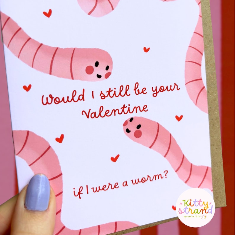 worm Valentine's Day card. pink Valentine's day card with an illustration of two worms surrounded by love hearts and text reading Would I still be your valentine if I were a worm? in red typography. Valentine's Day card for boyfriend, husband or partner held by a woman in front of a cosy background