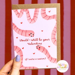worm Valentine's Day card. pink Valentine's day card with an illustration of two worms surrounded by love hearts and text reading Would I still be your valentine if I were a worm? in red typography. Valentine's Day card for boyfriend, husband or partner held by a woman in front of a cosy background