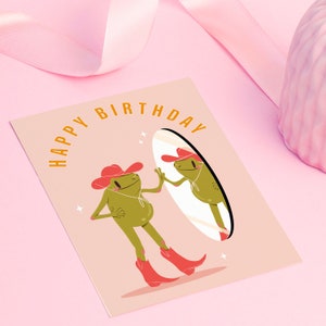 Pink and green cowboy frog happy birthday card. pink greetings card with a green frog in a cowboy hat and cowboy boots looking I the mirror and smiling. text on the card reads happy birthday in yellow lettering. Card rests against a pink background