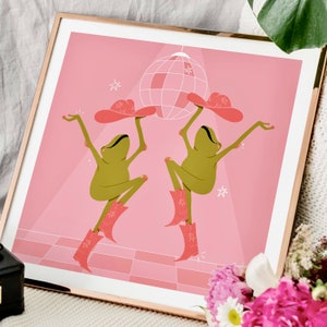 Gold frame leans against white sofa surrounded by flowers. Art in frame features a UPA style frog illustration. Two Green smiling frogs dance beneath a sparkling disco ball wearing pink cowgirl boots and a pink cowgirl hat