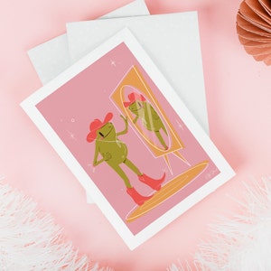 pink and green cowboy frog art print of a cartoon style frog wearing a pink cowgirl hat and pink cowboy boots looking into a vintage yellow mirror. Print features a soft pink background and rests on a soft pink table