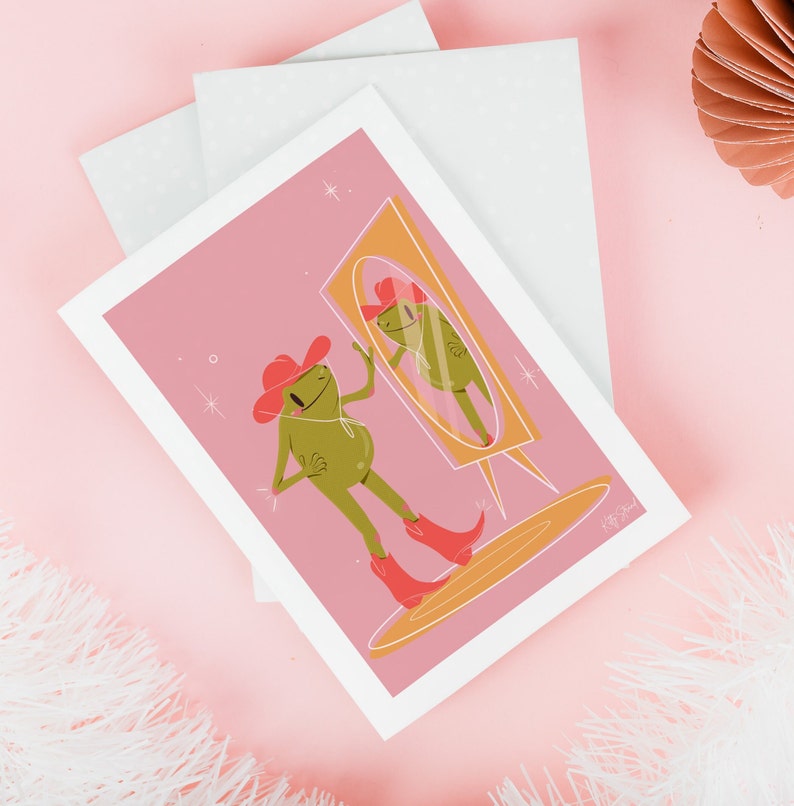 pink and green cowboy frog art print of a cartoon style frog wearing a pink cowgirl hat and pink cowboy boots looking into a vintage yellow mirror. Print features a soft pink background and rests on a soft pink table