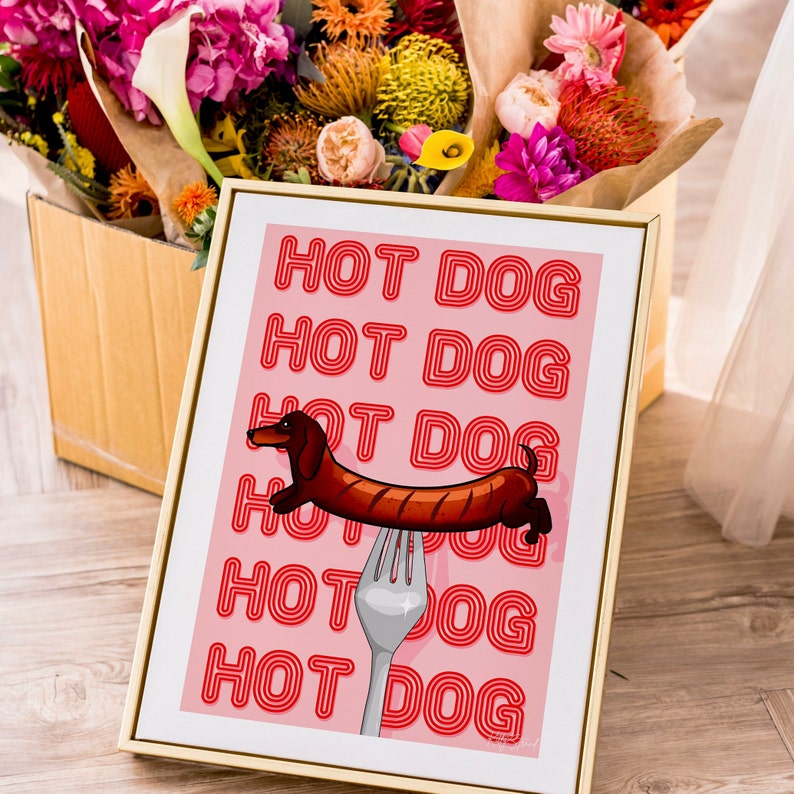 Printable pink & red dog art print. Soft pink background with red text in the style of neon lights reading HOT DOG. in the centre of the art print is a dachshund in the shape of a sausage on the end of a silver fork in a playful style in gold frame