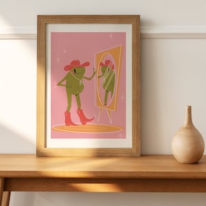 On a retro sideboard a wooden frame and green house plant sit. Art print in the frame features a UPA style frog illustration. A Green smiling frog looks into a tall yellow mirror wearing pink cowgirl boots and a pink cowgirl hat