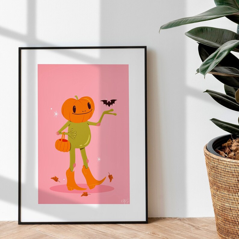 howdy halloween frog art print in pink, green and orange next to houseplant. UPS style cartoon illustration of a frog with a pumpkin head wearing orange cowboy boots and carrying a pumpkin bag. next to him flies a little bat, leaves surround his feet