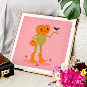 square howdy halloween frog art print in pink, green and orange. UPS style cartoon illustration of a frog with a pumpkin head wearing orange cowboy boots and carrying a pumpkin bag. next to him flies a little bat, leaves surround his feet. gold frame