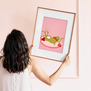women holds gold frame against a pink living room wall. in the frame is a pink and red frog aesthetic art print