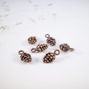 Könnte beinhalten: Nahaufnahme von mehreren kleinen, bronzefarbenen Tannenzapfen-Anhängern. Jeder Anhänger hat eine strukturierte Oberfläche und eine kleine Schlaufe oben zum Befestigen. Die Anhänger sind auf einer weißen Oberfläche verstreut.