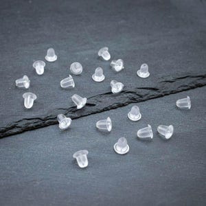 Könnte beinhalten: Eine Ansammlung klarer, durchscheinender Ohrringverschlüsse auf einer dunkelgrauen Oberfläche. Die Ohrringverschlüsse sind klein, mit einem abgerundeten Boden und einem Loch für den Stift. Die Gesamtästhetik ist schlicht und funktional.