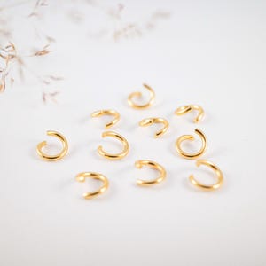 Könnte beinhalten: Eine Ansammlung kleiner, goldfarbener, offener Schmuckringe, die auf einer weißen Oberfläche verstreut sind. Die Ringe sind gleichmäßig verteilt und scheinen aus Metall zu bestehen. Der Hintergrund ist ein weiches Weiß mit einem Hauch von floralen Elementen.