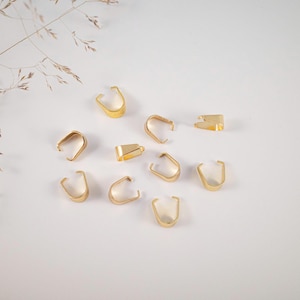 Könnte beinhalten: Eine Sammlung goldfarbener Schmuckbügel, präsentiert auf einer weißen Oberfläche. Die Bügel haben verschiedene Formen, darunter U-förmige und rechteckige Designs. Das Bild ist gut beleuchtet und hebt den metallischen Glanz der Bügel hervor.