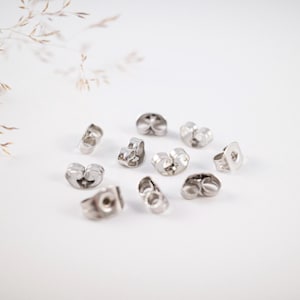 Könnte beinhalten: Eine Ansammlung silberfarbener Ohrringverschlüsse auf einer weißen Oberfläche. Die Ohrringverschlüsse sind klein und haben ein Schmetterlingsdesign. Das Bild ist gut beleuchtet und hebt den metallischen Glanz der Ohrringkomponenten hervor.