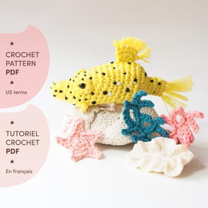 Peut inclure: Divers éléments crochetés à la main, dont un poisson jaune à pois noirs, une structure blanche en forme de corail et diverses créatures marines roses et turquoise. L'image contient également le texte "CROCHET PATTERN PDF" et "En français".
