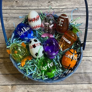 May include: A blue basket filled with colorful plastic Easter eggs decorated with sports themes and names. The eggs are decorated with baseball, football, basketball, soccer, and other sports designs. The basket is filled with green and blue shredded paper.