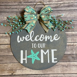 May include: A round, gray wooden sign with the words "welcome to our home" in white. A turquoise starfish replaces the "O" in "HOME". The sign is decorated with a bow featuring seashells and anchors, and turquoise berries.