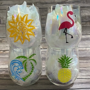 May include: Four iridescent glass wine glasses with different tropical designs. The designs include a yellow sun, a pink flamingo, a green palm tree with a blue wave, and a yellow pineapple.