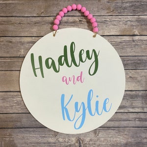 May include: A circular white wooden sign featuring the text Hadley in green, and in pink, and Kylie in light blue. The sign is suspended by a jute rope adorned with a series of bright pink wooden beads, creating a decorative hanging wall plaque.