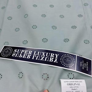 Op de afbeelding: Close-up van een lichtgroene stof met een herhalend bloemenpatroon. De stof heeft een geschulpte rand en een label met de tekst "SUPER LUXURY MAN LACE ORIGINAL". De stof is 147 cm breed.