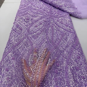 May include: Lavender beaded lace fabric with intricate patterns. The fabric is embellished with small, round beads and shimmering accents, creating a textured and luxurious appearance. The design features floral and geometric motifs.