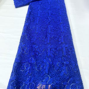 May include: A vibrant royal blue lace fabric featuring an intricate floral pattern with embroidered leaves and rose-like blossoms. The textile is embellished with small, shimmering sequins throughout the design, creating a textured and detailed surface.