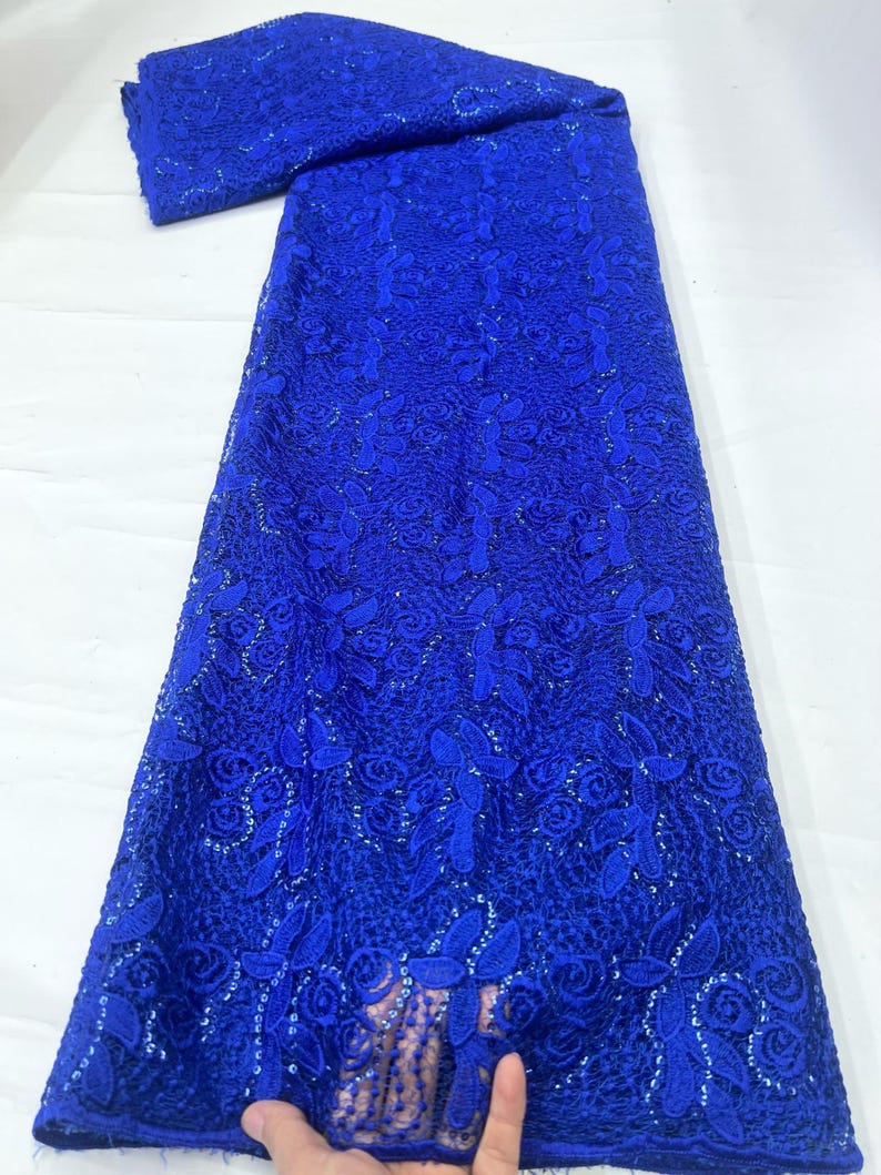 May include: A vibrant royal blue lace fabric featuring an intricate floral pattern with embroidered leaves and rose-like blossoms. The textile is embellished with small, shimmering sequins throughout the design, creating a textured and detailed surface.