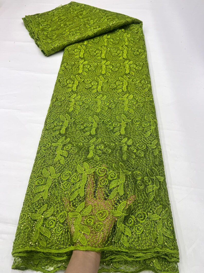 May include: A vibrant lime green lace fabric featuring an intricate embroidered pattern of swirling floral motifs and leaves. The textile has a delicate mesh base with a subtle shimmering finish throughout, showcasing a repeating botanical design.