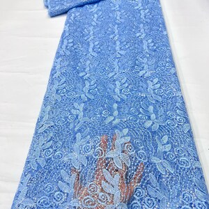 May include: A light blue lace fabric featuring an intricate floral pattern with embroidered leaves and swirling designs. The material is embellished with small, shimmering sequins throughout the mesh base, creating a textured and decorative appearance.