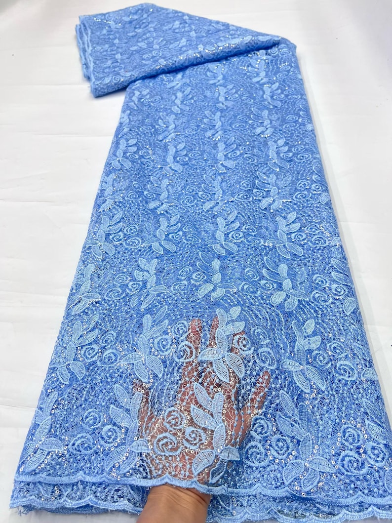 May include: A light blue lace fabric featuring an intricate floral pattern with embroidered leaves and swirling designs. The material is embellished with small, shimmering sequins throughout the mesh base, creating a textured and decorative appearance.
