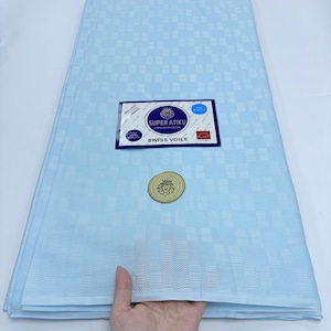 May include: A light blue fabric featuring a subtle, repeating geometric checkered pattern. A rectangular label is attached to the center, displaying the text Super Atiku 100% Egypt Cotton Swiss Voile, along with a circular gold-colored emblem.