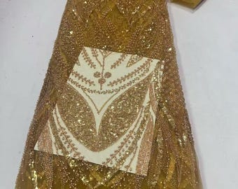 May include: Mustard yellow fabric with intricate gold bead and sequin embellishments. A cream-colored square with gold leaf-like designs is placed on top of the fabric. The fabric is likely intended for formal wear or special occasion garments.
