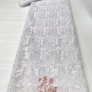 May include: A white lace fabric featuring an intricate floral pattern with embroidered roses and leaves. The material has a sheer, mesh-like base with a delicate texture and a subtle shimmer throughout the design, presented in a long, folded bolt.