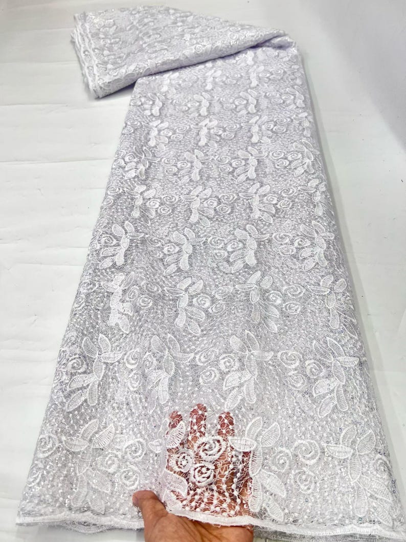 May include: A white lace fabric featuring an intricate floral pattern with embroidered roses and leaves. The material has a sheer, mesh-like base with a delicate texture and a subtle shimmer throughout the design, presented in a long, folded bolt.