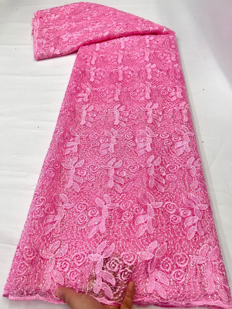 May include: A vibrant pink lace fabric featuring an intricate floral pattern with embroidered leaves and rose-like blossoms. The textile is embellished with small, shimmering sequins throughout the mesh base, creating a textured and reflective surface.