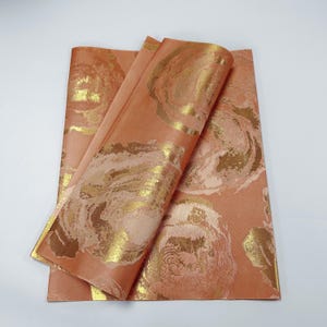 Puede incluir: Dos hojas de papel de regalo de color melocotón con un patrón floral dorado. El papel está doblado por la mitad y el patrón es visible en ambos lados.