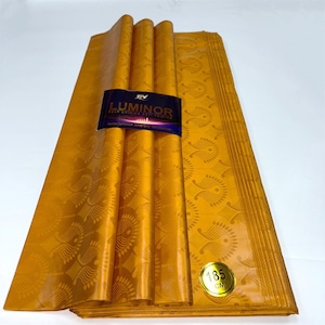 May include: A stack of gold fabric with a repeating pattern of stylized leaves. The fabric is labeled "Luminor Deep Shining Anti-Fading" and "185 cm".
